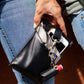 A person holding  a black vegan leather phone pouch with a black and grey vegan leather magnetic wristlet key chain and a black and gray removable three pocket wallet and black lip balm holder set. 